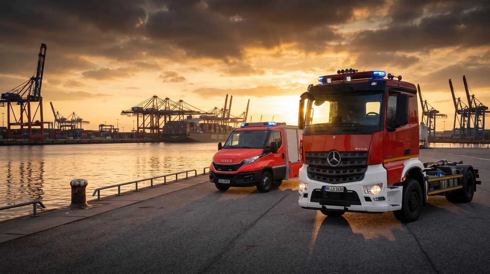 Ein rotes Feuerwehrfahrzeug auf IVECO-Basis und ein rot-weißer Mercedes-Benz Arocs mit Abrollaufbau stehen bei Sonnenuntergang an einer Hafenkante, im Hintergrund Containerbrücken.